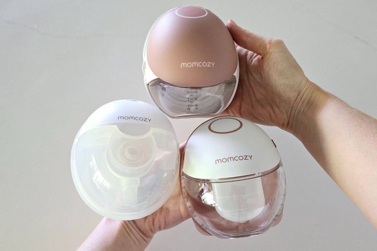 A person holding three Momcozy wearable breast pumps—the Momcozy M5, M6, and M9—against a neutral background. The pumps have a sleek, compact design with different color accents: the M5 in white, the M6 in soft pink, and the M9 in white with rose gold detailing. The transparent milk collection cups show volume markings, highlighting their storage capacity.