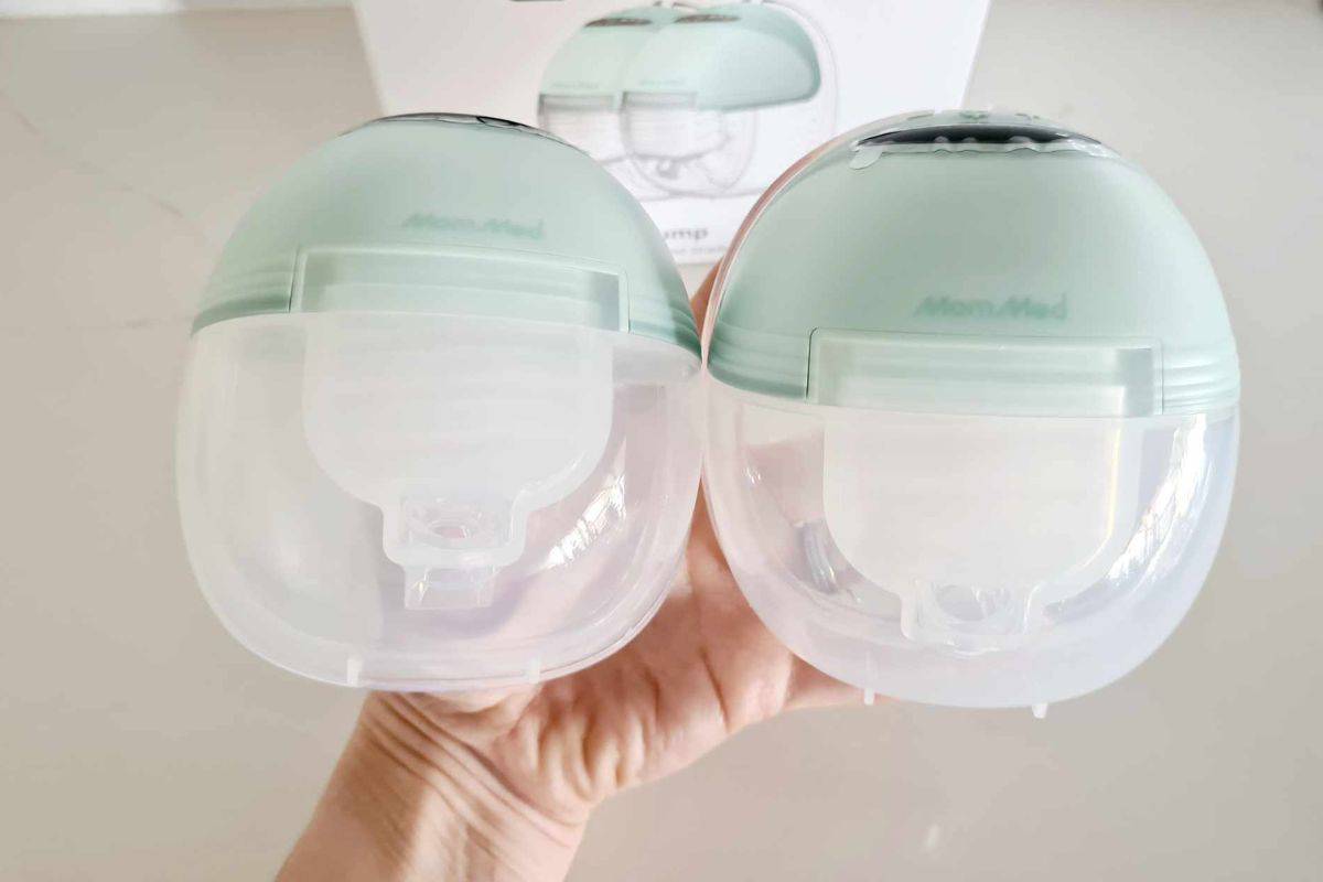 A close-up image of a person holding two MomMed S21 wearable breast pumps side by side. The pumps have a transparent milk collection container and a light green motor unit on top. The MomMed logo is visible on the motor casing, and the clear design allows for easy visibility of the pump's internal components. In the background, there is a slightly blurred image of the product packaging.