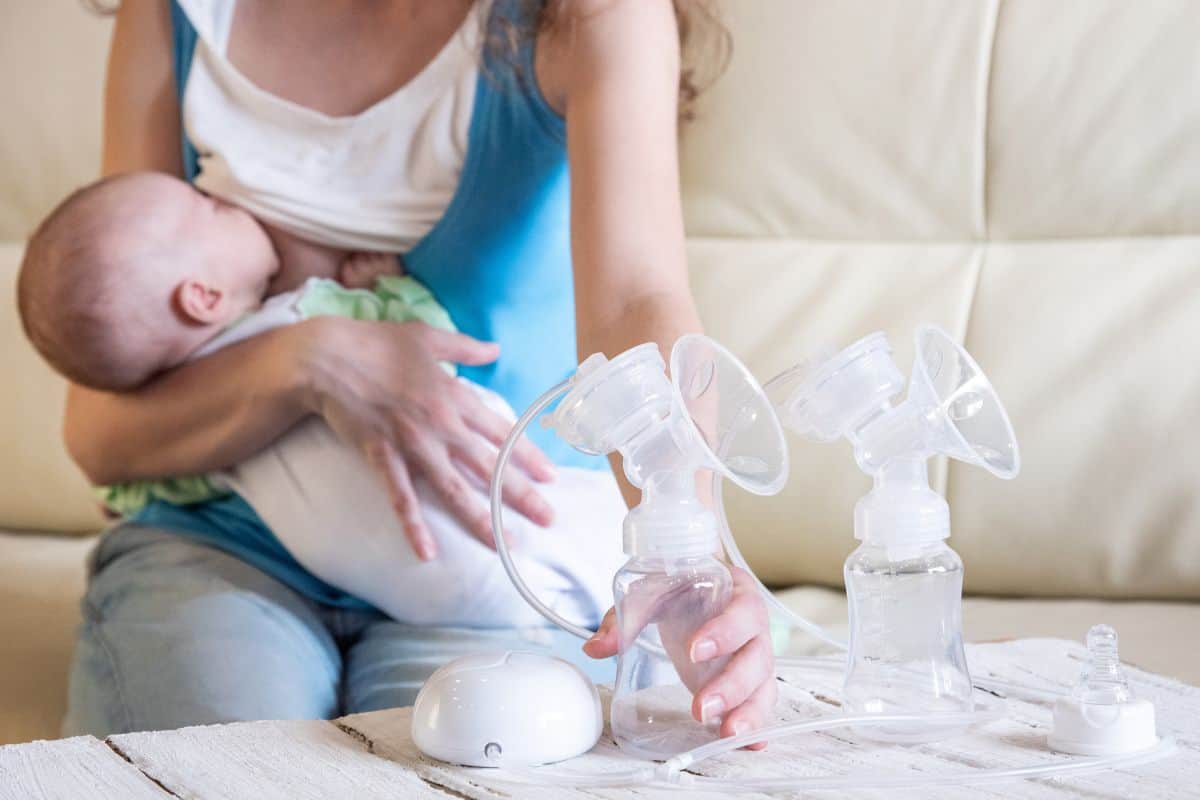 Mother breastfeeding baby while holding a double electric breast pump on a table—illustrating how to combine breastfeeding and pumping.