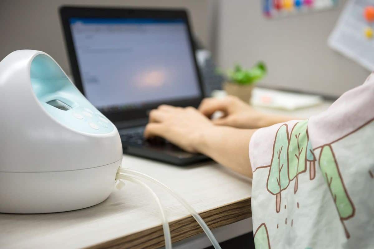 Working mom typing on a laptop at her desk while using an electric breast pump during a pumping break at work.