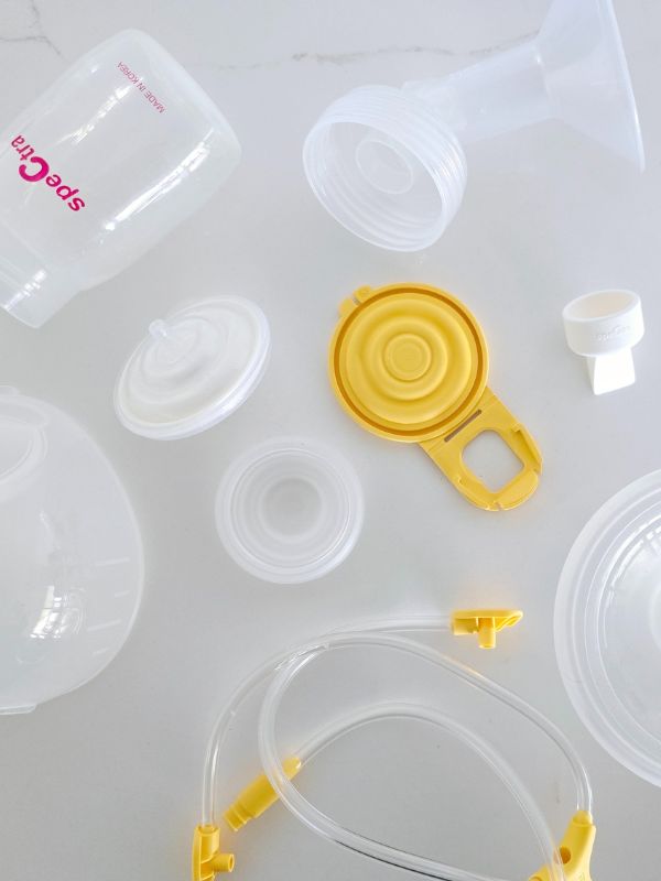 Flat lay of assorted breast pump accessories from the Spectra and Medela brands, including flanges, tubing, valves, bottle caps, membranes, and connectors, all arranged on a white surface.