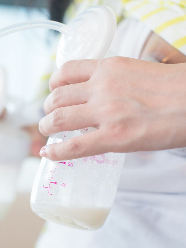 Close-up of a person holding a breast pump bottle partially filled with breast milk during a pumping session. The flange is attached to the breast, and the milk is being collected into a marked bottle.