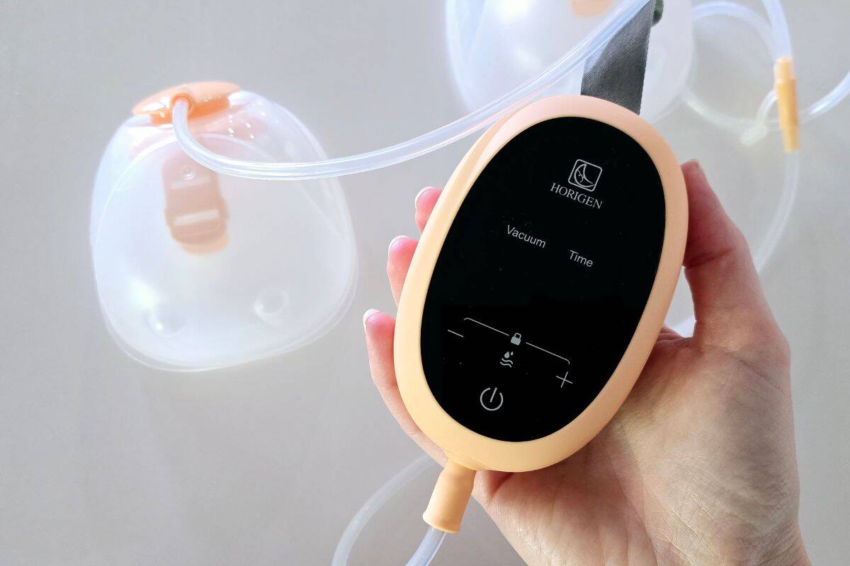 Hand holding the Horigen D6 breast pump motor with touchscreen controls, connected to collection cups via tubing