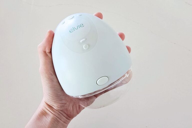 Hand holding the Elvie Pump, a sleek white wearable breast pump with a transparent 5oz collection bottle attached, shown against a light background.